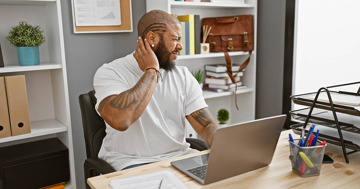 Man holding the back of his neck in pain while sitting at an office desk with a laptop, showing the strain of air conditioning on the spine