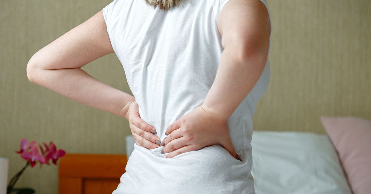Woman standing in her bedroom holding her lower back in pain after waking up, illustrating the morning stiffness characteristic of ankylosing spondylitis