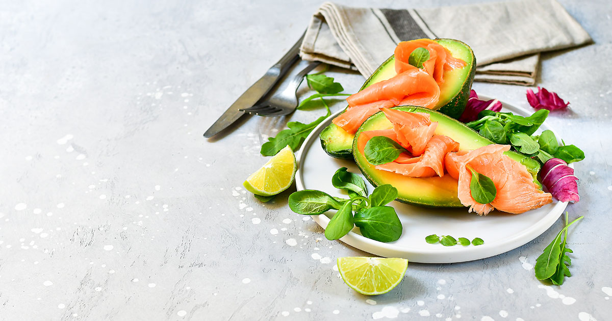 A plate of smoked salmon over halved avocados with fresh arugula and lime wedges on a white plate, representing an anti-inflammatory meal to reduce spinal inflammation