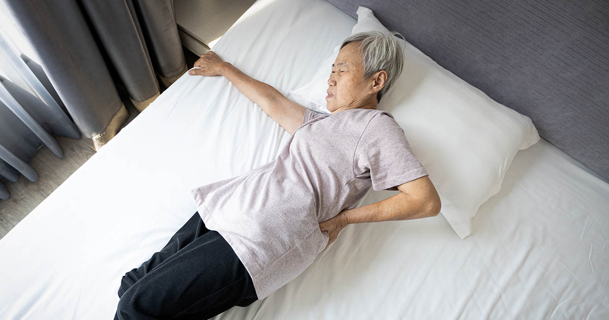 Elderly woman lying in bed pressing her hand against her lower back in pain, illustrating nocturnal back pain that worsens at night