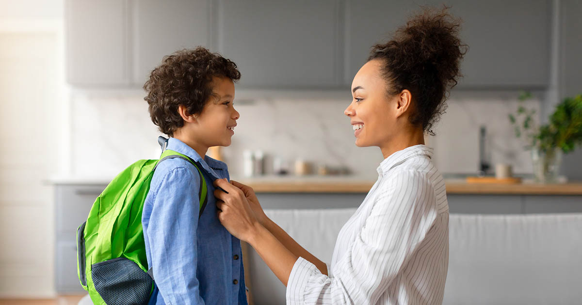 Mother kneeling to adjust the green backpack straps on her young son before school, ensuring correct fit to protect his spine