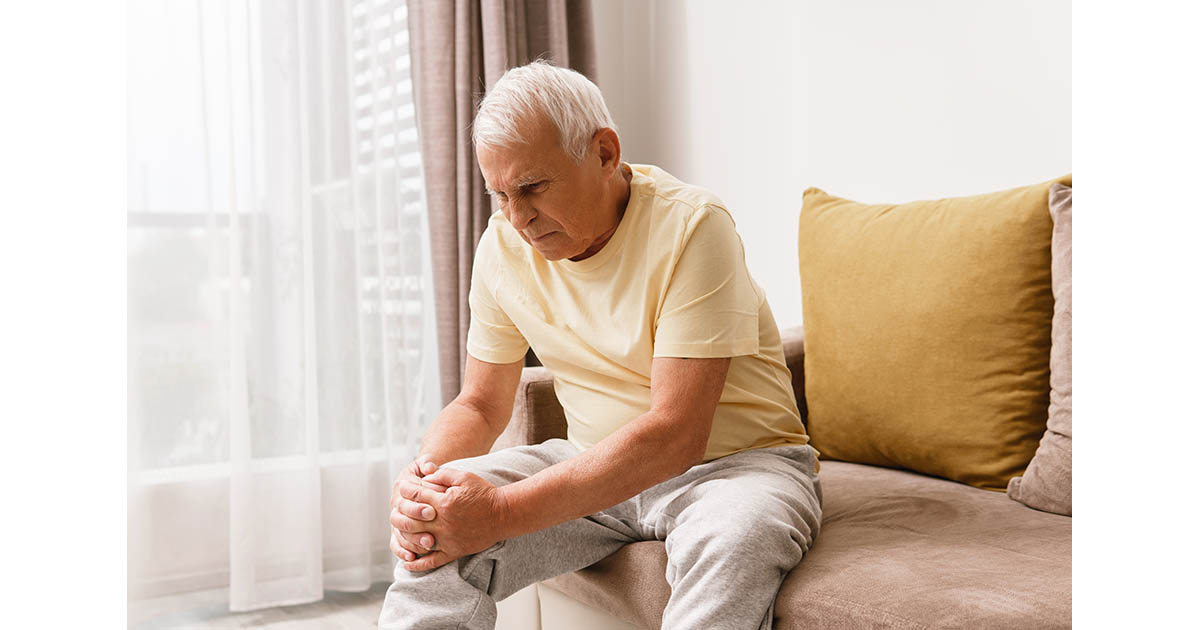 Senior man sitting on a couch at home holding his knee in pain, with soft window light behind him suggesting overcast or rainy conditions