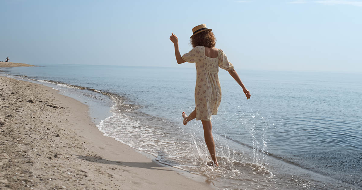 Woman walking barefoot through shallow ocean waves on a sandy Florida beach, arms out for balance as she kicks water