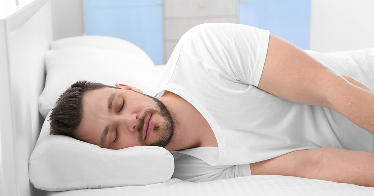 Man lying on his side in bed, sleeping on a contoured orthopedic cervical pillow that supports the natural curve of the neck