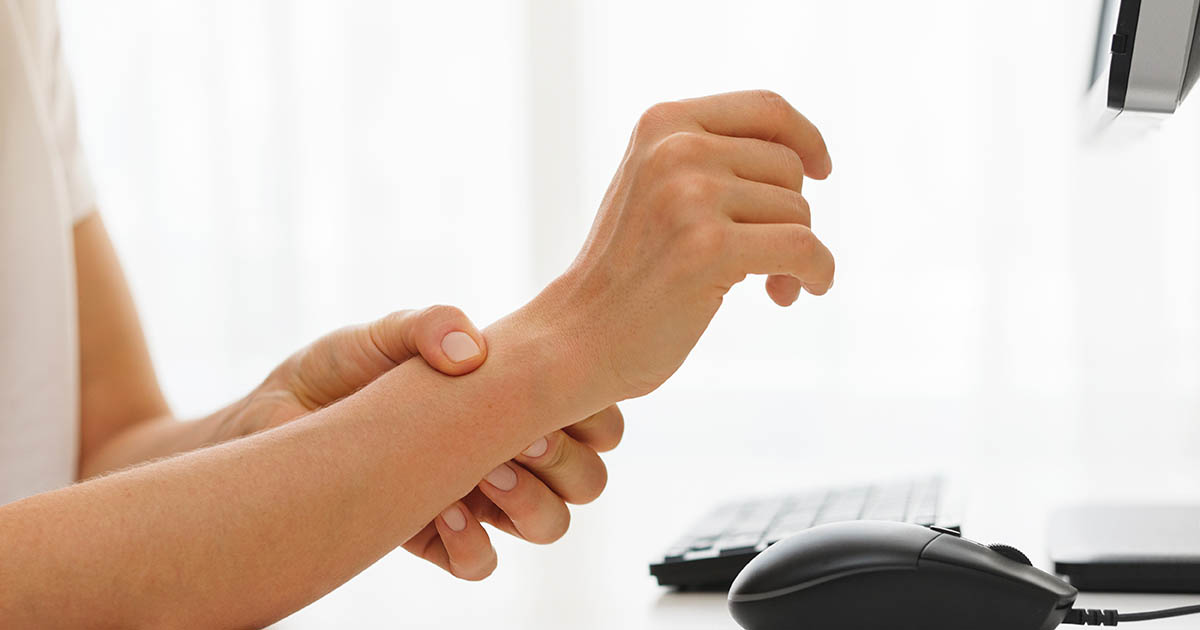 Person seated at a computer desk gripping their wrist in pain, illustrating the hand and wrist discomfort pattern of carpal tunnel syndrome