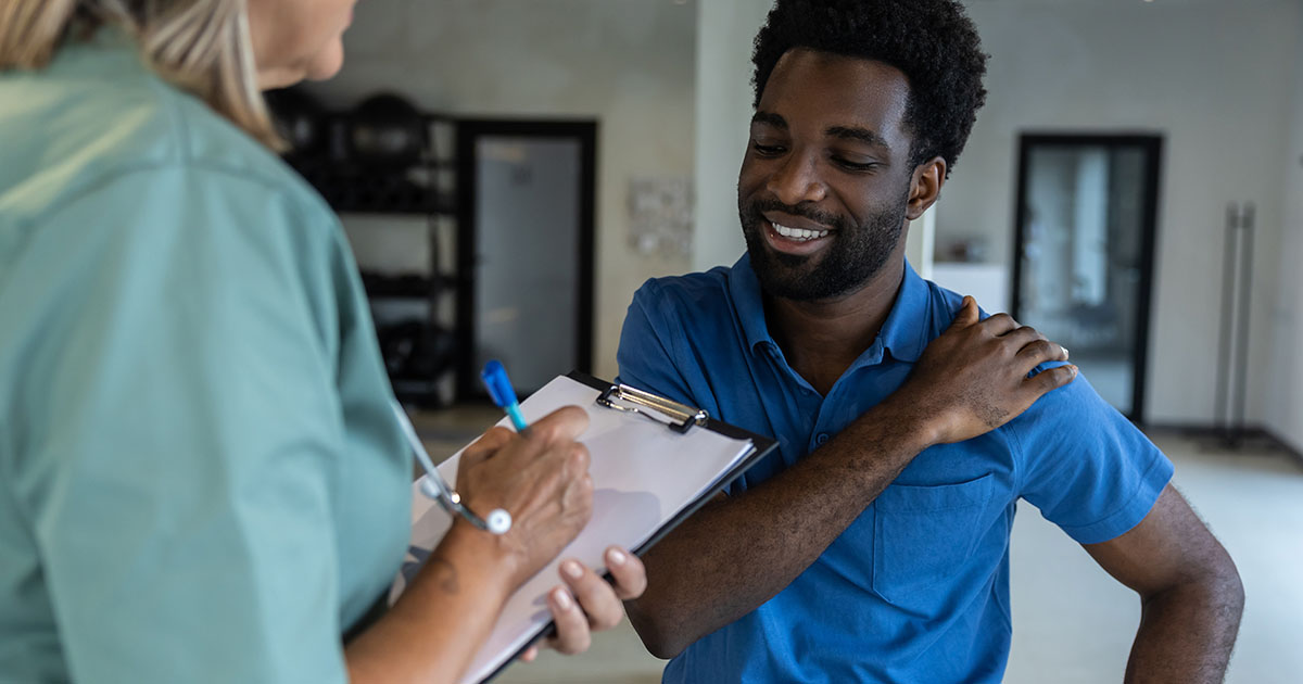 Physical therapist in teal scrubs evaluating a male patient's shoulder range of motion while taking notes on a clipboard in a clinical rehabilitation setting