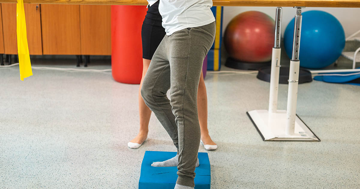 Patient performing a single-leg balance exercise on a stability board in a physical therapy clinic, with a therapist providing guidance
