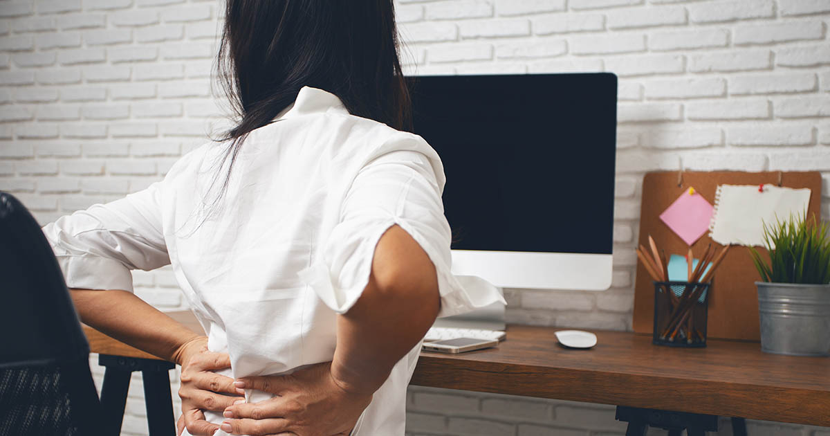Woman sitting at office desk with both hands pressed into her lower back, showing pain from prolonged sitting