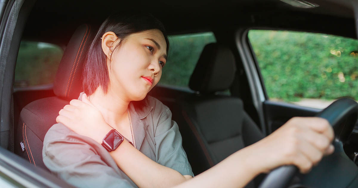 Woman in a car reaching behind her head to grip her neck and shoulder in pain, with a red pain indicator overlay, illustrating the discomfort of driving long hours on the highway