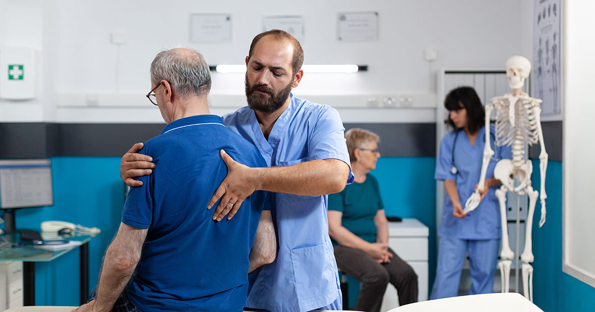 Chiropractor in medical scrubs working on an older patient's upper back in a clinical setting, with a spinal anatomy model visible in the background