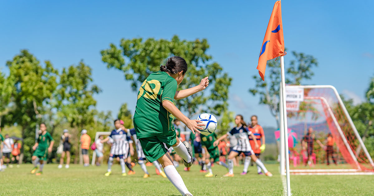 Male athlete in black tank top seen from behind with both hands pressed into his lower back in pain, standing outdoors with green trees behind him, illustrating back pain from sports activity