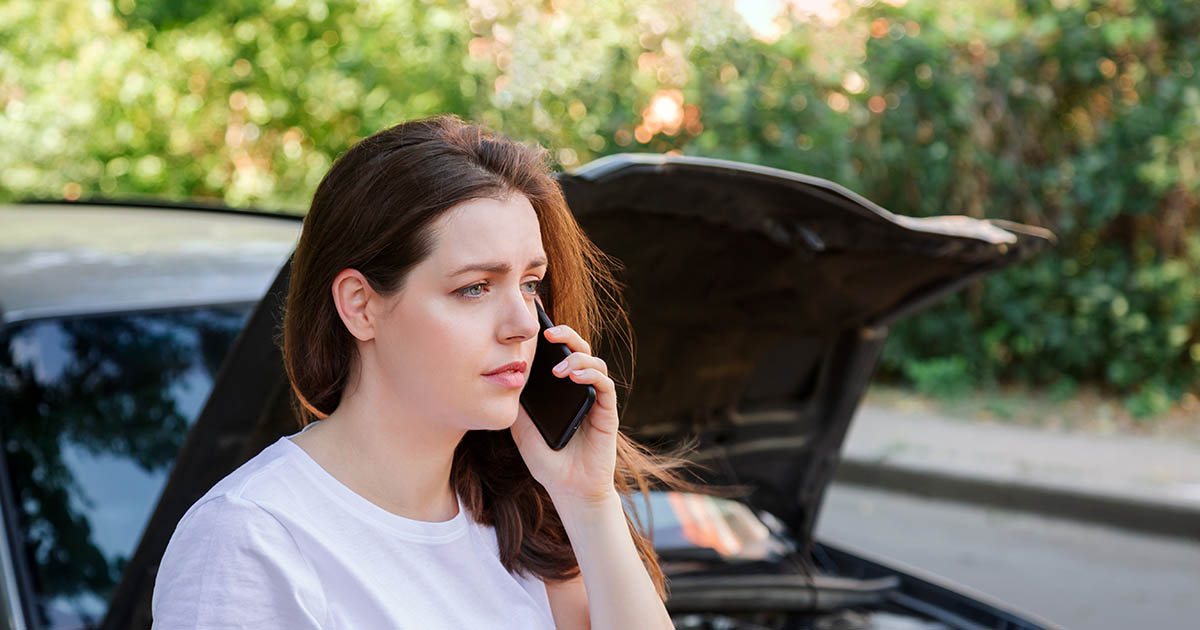 Young woman in a white t-shirt standing beside a car with its hood raised, talking on her phone with a worried expression after a car accident