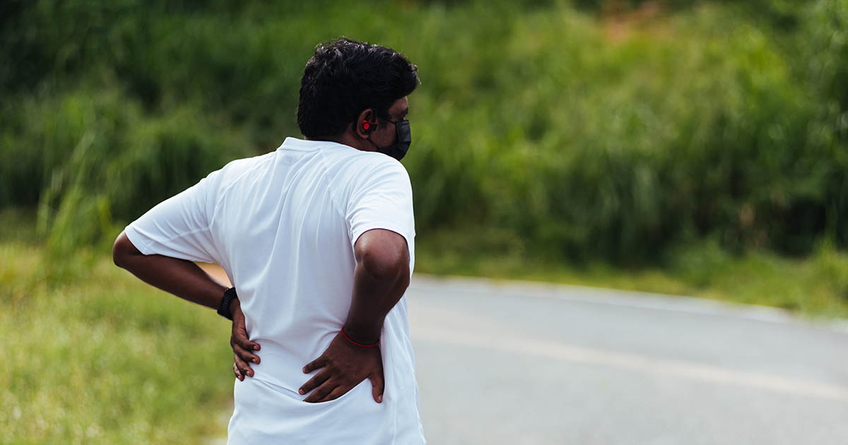 Man in white shirt gripping his lower back in pain while stopped on an outdoor road, with lush green trees visible in the background