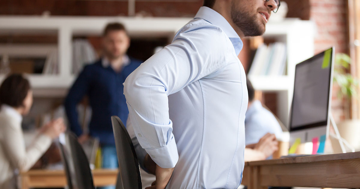 Businessman pressing his fist into his lower back while seated at a desk in a shared office, illustrating how prolonged sitting leads to glute inhibition and chronic back pain
