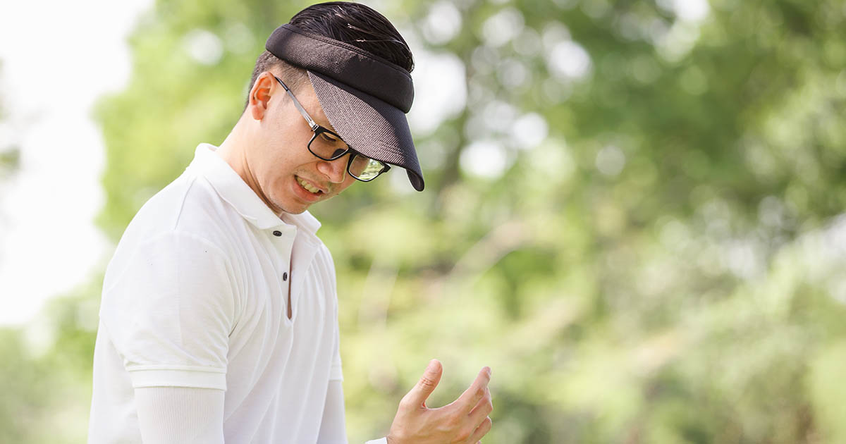 Golfer wearing a visor and glasses wincing in pain on an outdoor golf course with green trees in the background, illustrating golf-related back and spine injury