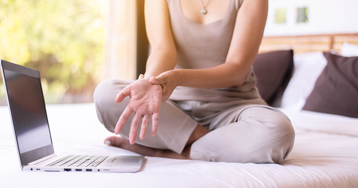 Woman sitting on the edge of her bed in soft morning light, holding her wrist as she examines a tingling, numb palm
