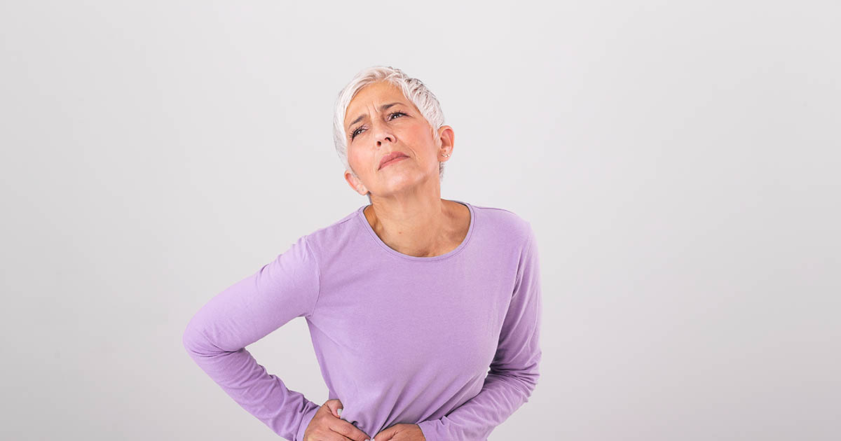 Older woman in a purple sweater wincing and pressing both hands into her lower back and hip, showing the classic pain location for hip and sciatic-pattern complaints