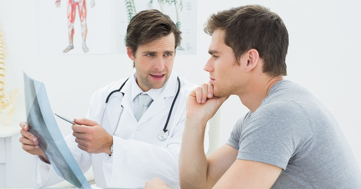 Male chiropractor reviewing spine X-ray with a male patient in a clinical consultation room, discussing a treatment plan