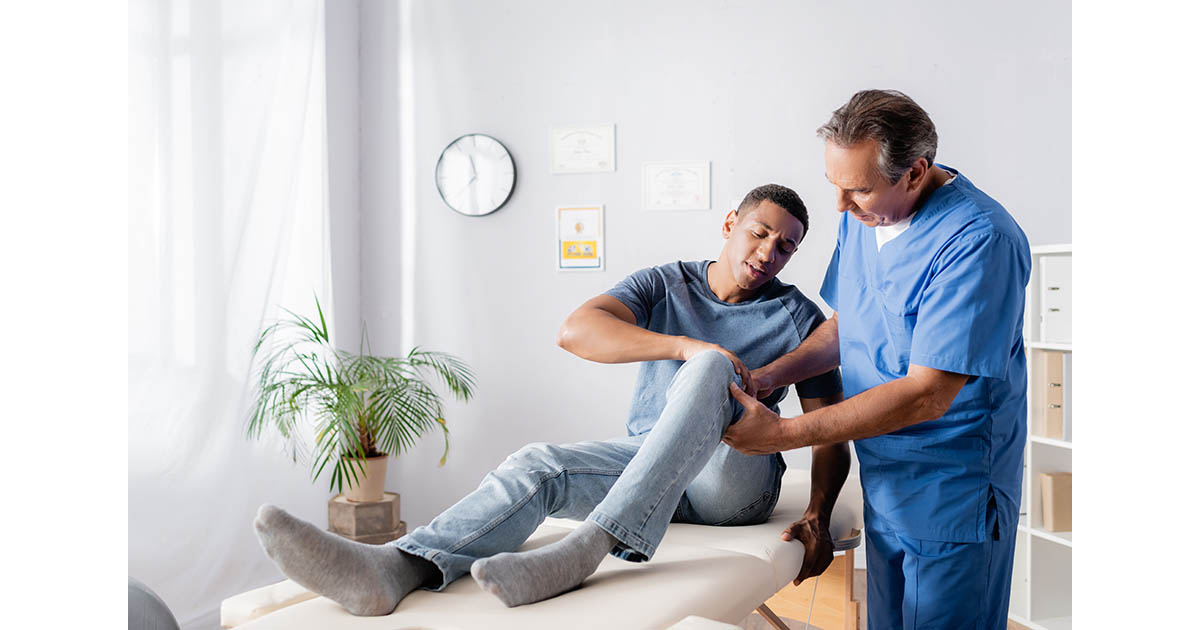 Chiropractor in blue scrubs examining and palpating the knee of a young male patient lying on a treatment table in a bright clinical room