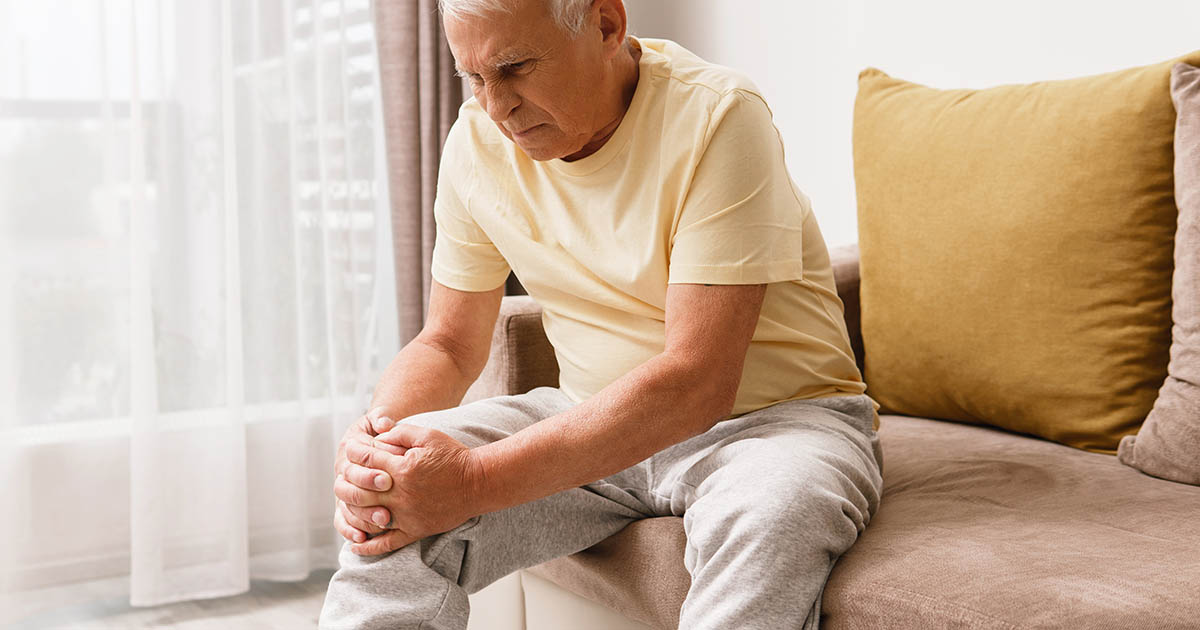 Senior man sitting on his couch at home, both hands wrapped around his knee, demonstrating the unexplained knee pain that often develops without a fall or injury