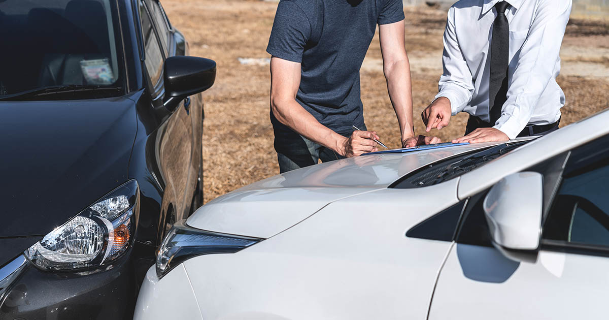 Two men standing beside damaged vehicles at a car accident scene, one signing a claim document on the hood of a white car