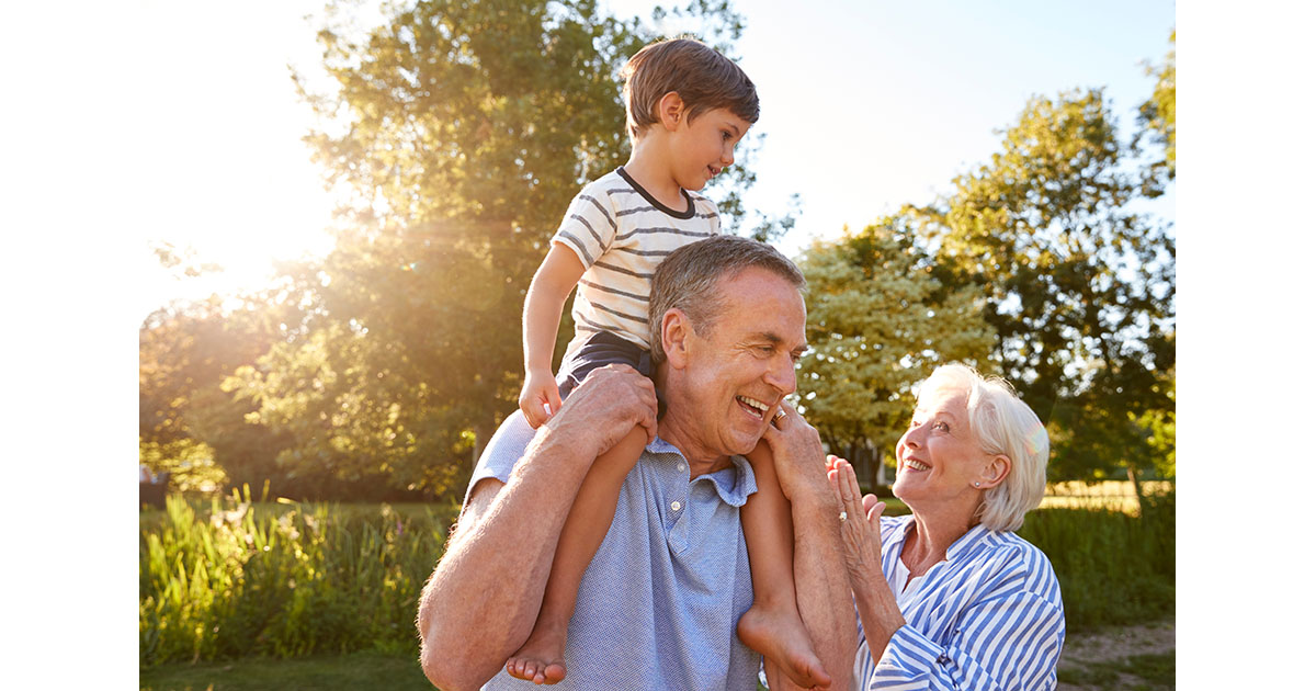 Grandparents lifting a young grandchild onto shoulders in a sunny park, all three laughing outdoors
