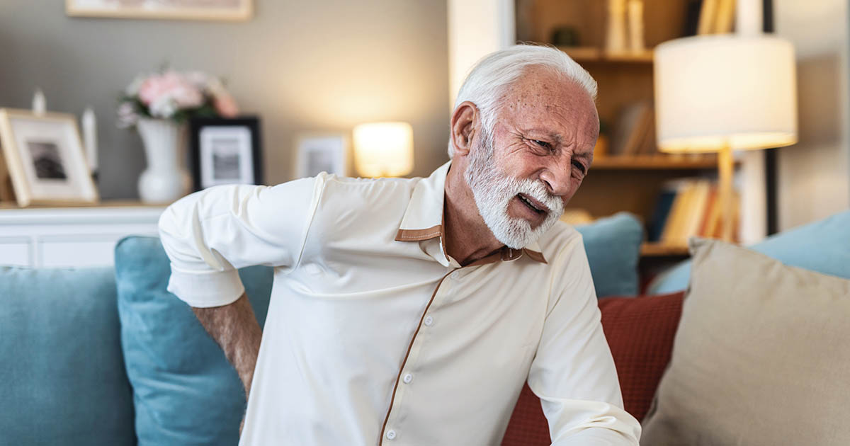 Older man in a white shirt sitting on a couch, grimacing and pressing his hand against his lower back in pain after a day of lifting and carrying grandchildren