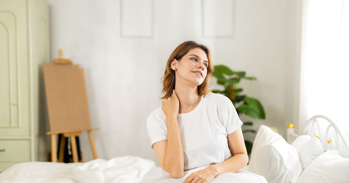 Woman sitting upright on her bed in morning light, holding her neck with a pained expression as she wakes with stiffness