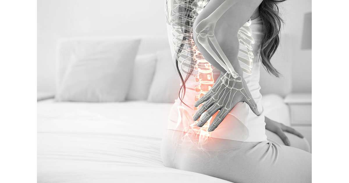 Woman sitting on the edge of a bed, holding her lower back, with a digital spine overlay highlighting the lumbar vertebrae in orange