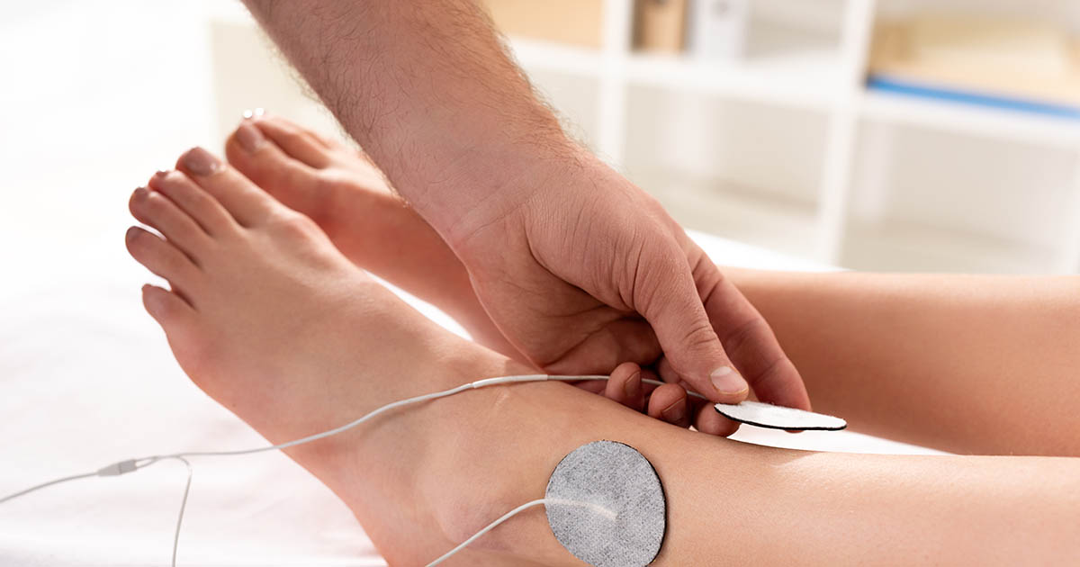 Clinician placing an electrode therapy pad on a patient's lower leg in a bright clinical treatment room, illustrating the electrotherapy component of a neuropathy recovery program