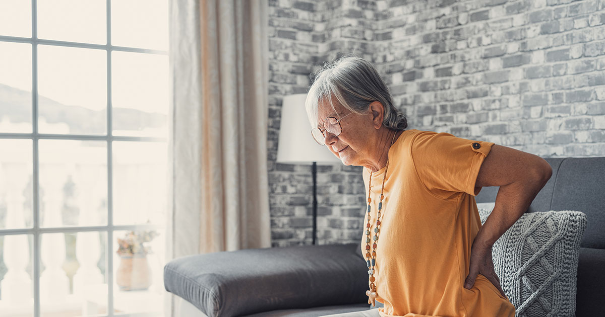 Older woman in an orange shirt sitting on the edge of a couch, both hands pressed against her lower back, showing the location of chronic back pain