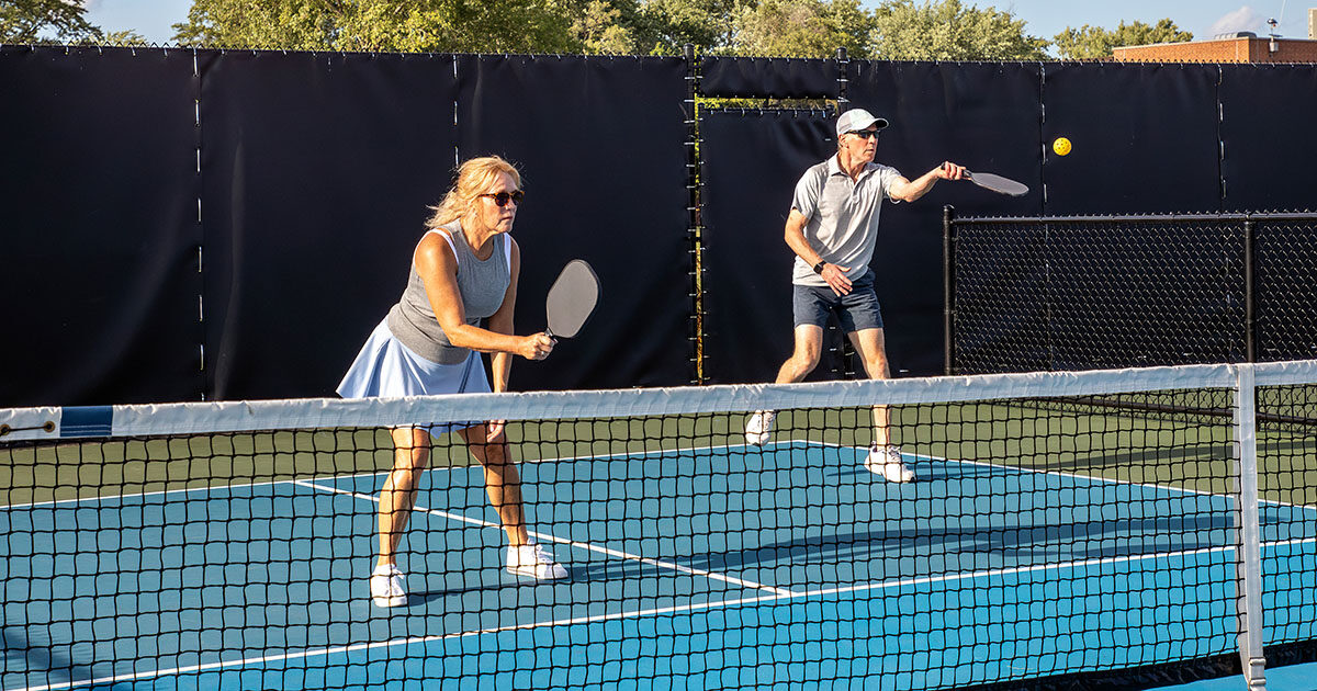 Two pickleball players, a woman and a man, competing on an outdoor court in afternoon sunlight, demonstrating the lateral movement and overhead swings that drive common pickleball injuries