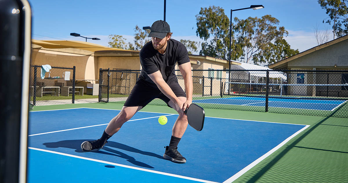 Pickleball player in black lunging low across a blue outdoor court to return a yellow ball with his paddle, the exact rotational stress that drives most shoulder, knee, and lower-back injuries the article covers