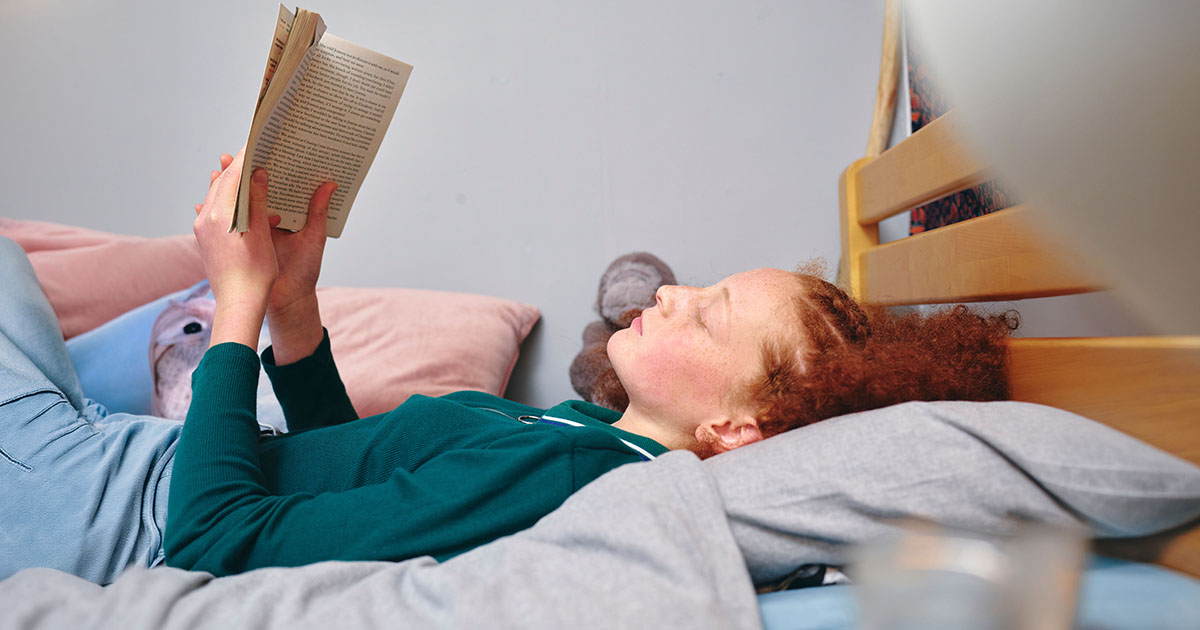 Person propped up on pillows in bed reading a book, the sustained cervical flexion position that drives neck stiffness and upper back tension