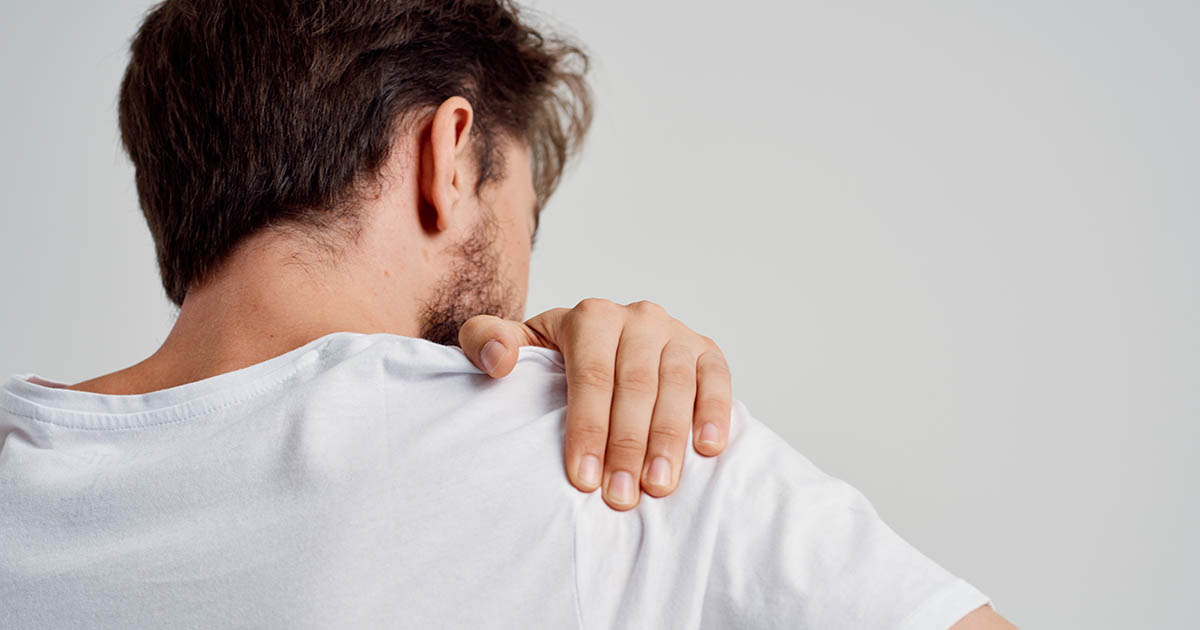 Man in a white t-shirt viewed from behind, reaching across to grip his painful shoulder, illustrating the classic location of rotator cuff pain