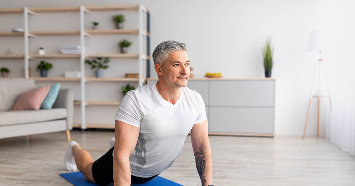 Middle-aged man in a white t-shirt doing a cobra press-up stretch on a blue yoga mat in his living room, performing a home exercise that helps relieve disc-based sciatica