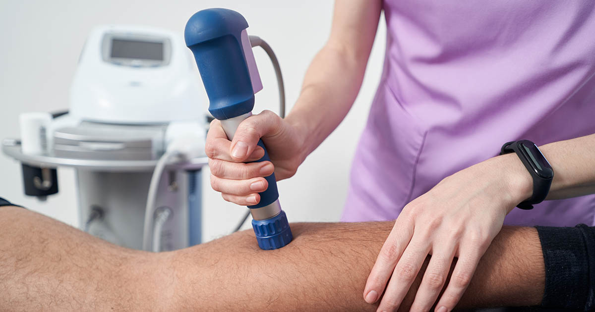Clinician in pink scrubs applying a blue shockwave therapy handpiece to a patient's leg in a clinical setting, with the shockwave device console visible in the background