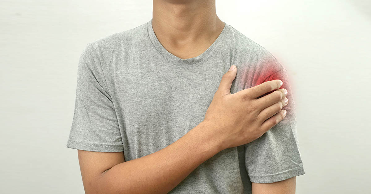 Man in a grey t-shirt pressing his hand against his shoulder with a red pain indicator overlay, illustrating the anterior shoulder pain location of subacromial impingement syndrome