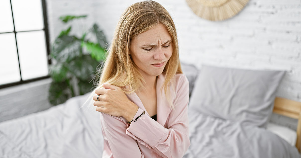 Woman in pajamas sitting on the edge of her bed in the morning, gripping her shoulder with a pained expression