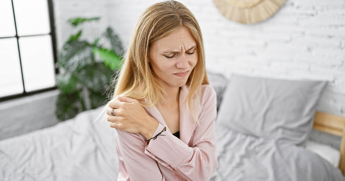 Young woman in pink pajamas sitting on the edge of her bed, wincing and gripping her shoulder in pain, white bedroom walls and disheveled sheets in the background