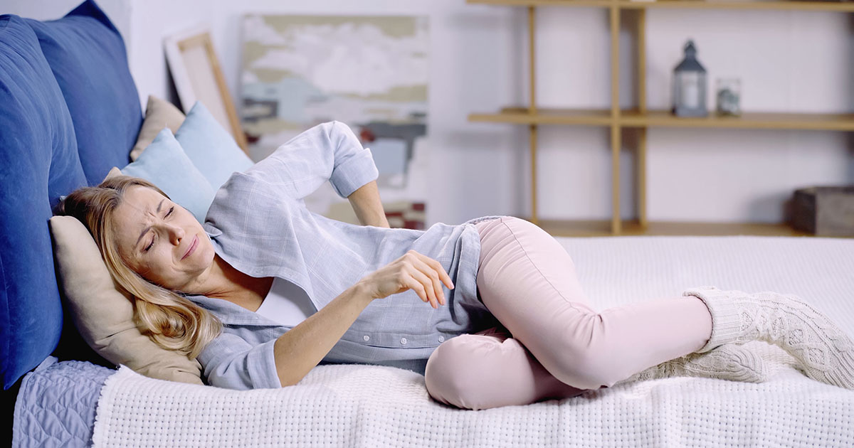 Woman lying on her bed in a bright bedroom, wincing in pain with her eyes closed and hand pressed against her lower back and hip, illustrating the sciatic nerve discomfort that disrupts sleep