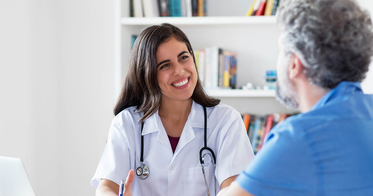 Smiling female doctor in white coat consulting warmly with a male patient in a bright clinic office, representing the bilingual Spanish-speaking care offered at Spine and Wellness Center Lakewood Ranch