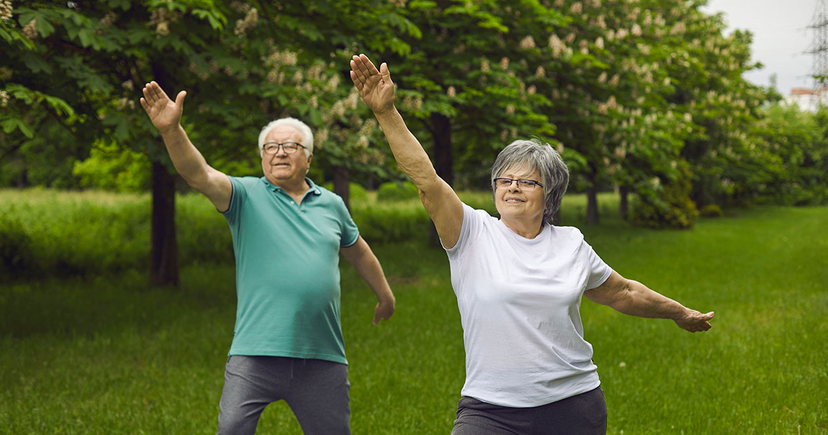 Older couple doing arm stretches outdoors in a park, smiling while exercising together to stay active and maintain mobility