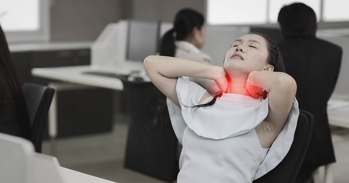 Woman at an office desk gripping her neck and upper back in pain, illustrating how chronic workplace stress physically loads the cervical and lumbar spine