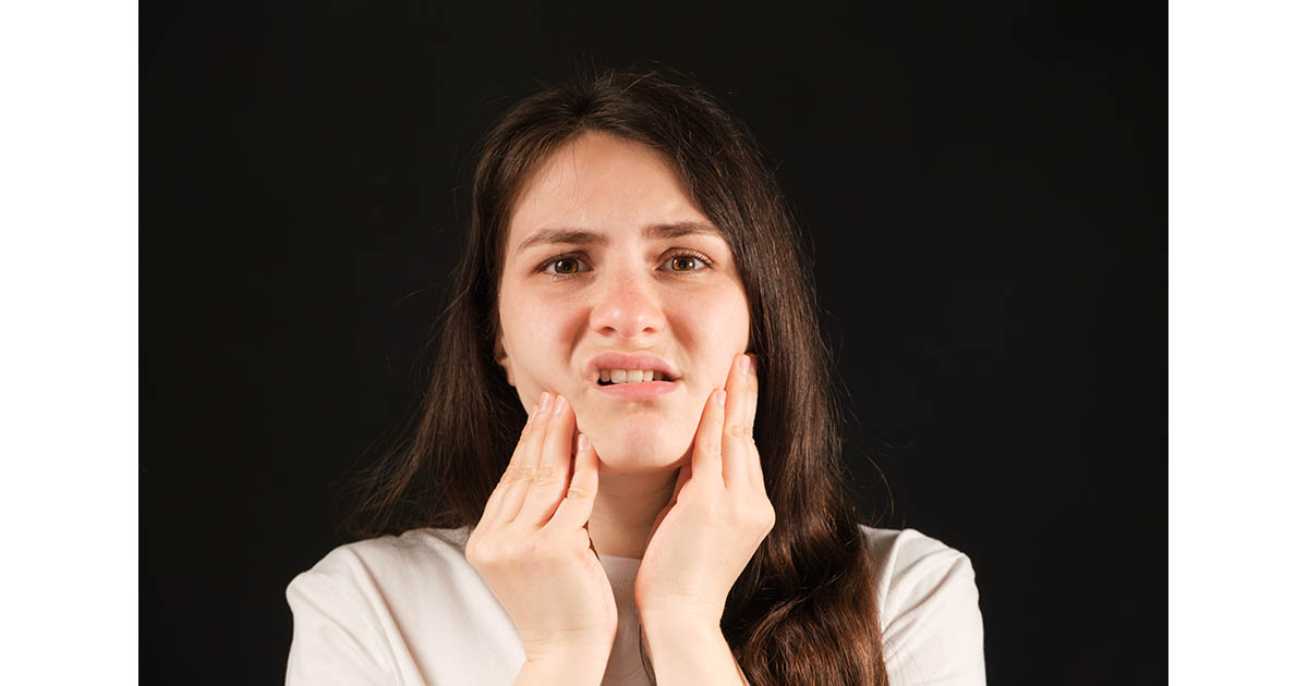 Woman pressing both hands to her jaw and cheeks, wincing in pain from temporomandibular joint dysfunction