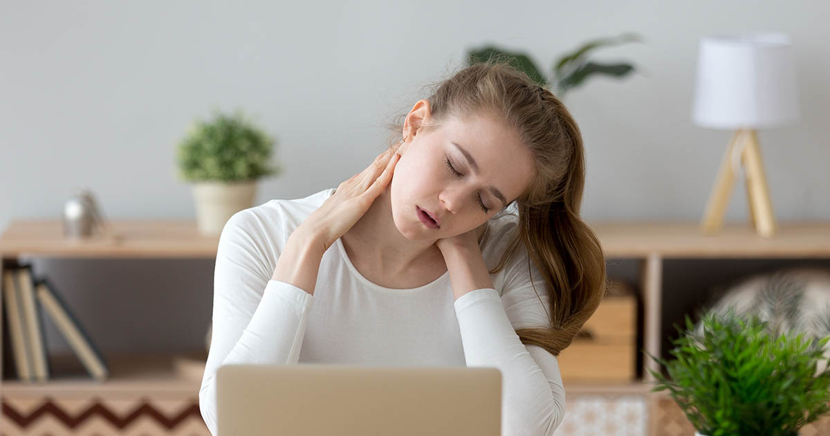 Young woman at her desk, eyes closed, pressing both hands to her neck and shoulder in pain, with a laptop open in front of her, illustrating the neck and shoulder discomfort pattern of upper cross syndrome