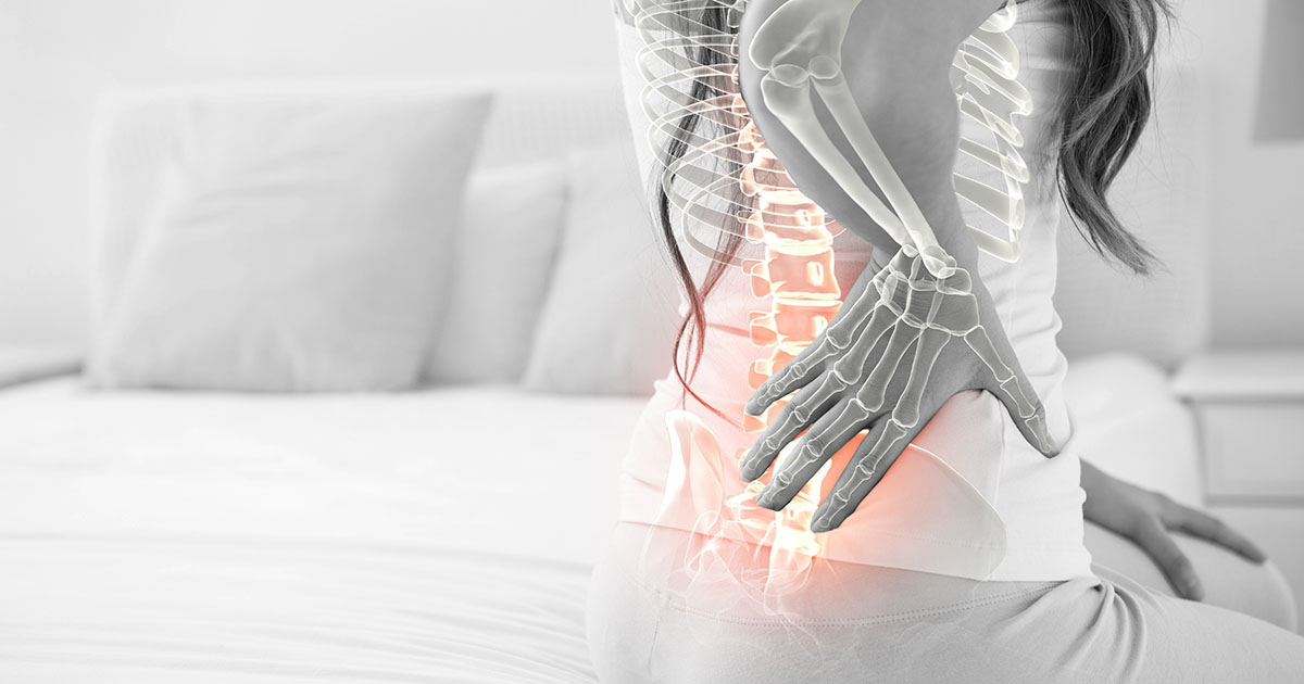 Woman sitting on a bed holding her lower back with a digital spine overlay highlighting lumbar pain area