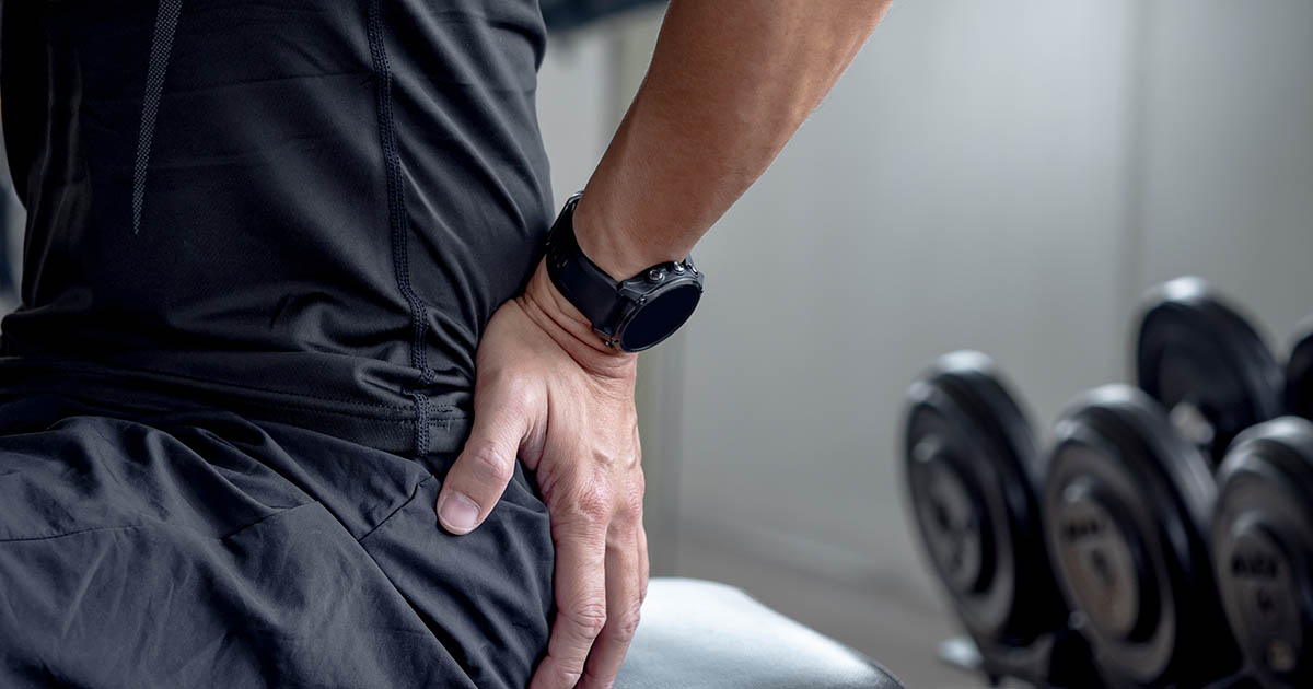 Man in gym attire seated and clutching his lower back in pain, with a rack of dumbbells visible in the background
