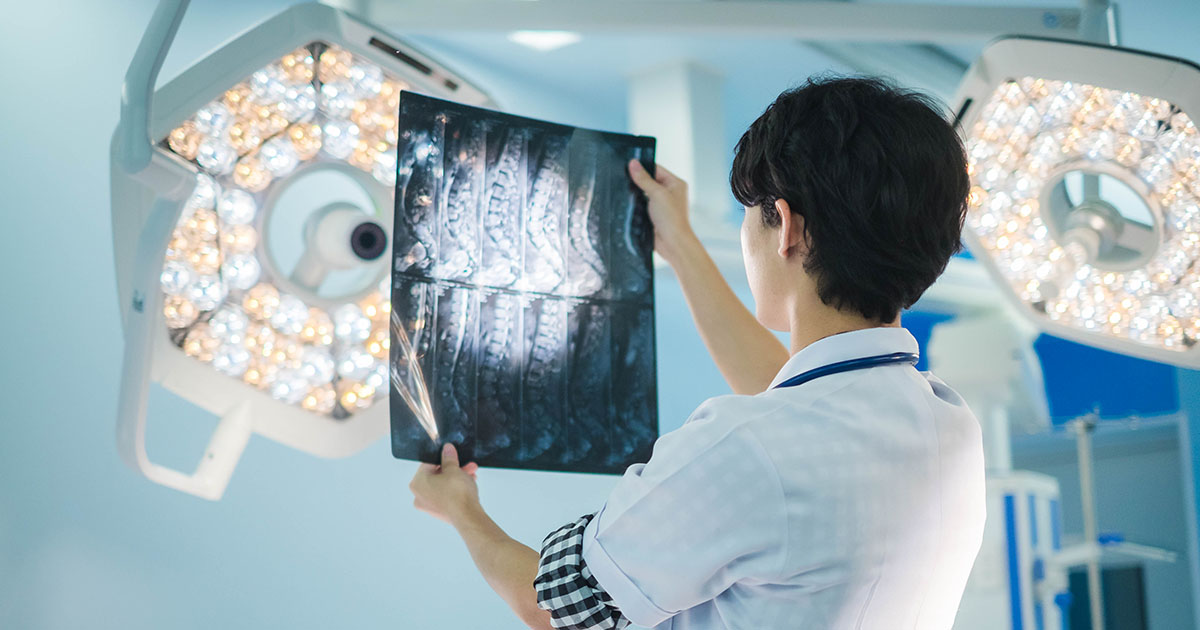 Doctor in a white coat reviewing a lumbar spine X-ray on a light box, pointing at vertebral structures to explain findings to a patient