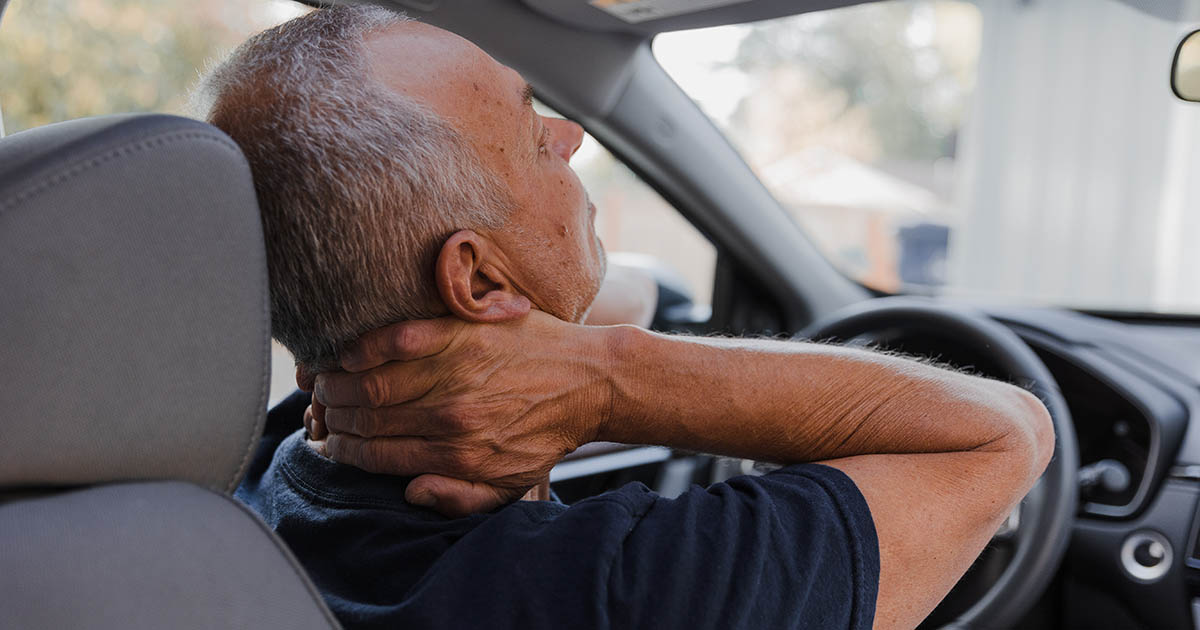 Middle-aged man sitting in a car, both hands gripping the back of his neck in pain, illustrating the persistent neck stiffness and discomfort that lingers weeks after a whiplash injury